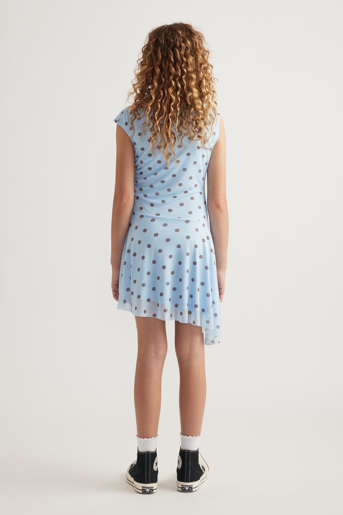Girl viewed from behind wearing a blue polka dot asymmetric dress with black sneakers