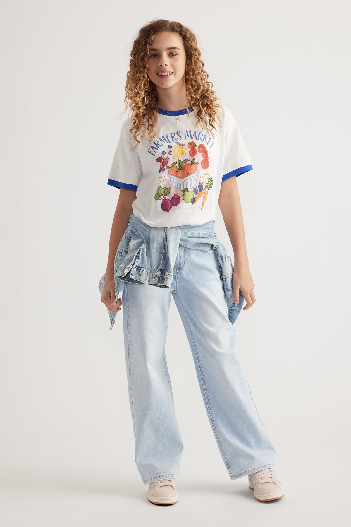 Girl wearing an oversized white graphic tee with blue trim, light wash wide leg jeans and a denim jacket tied at the waist