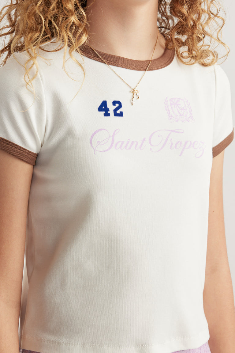 Girl wearing a cream ringer tee with brown contrast trim and a Saint Tropez graphic