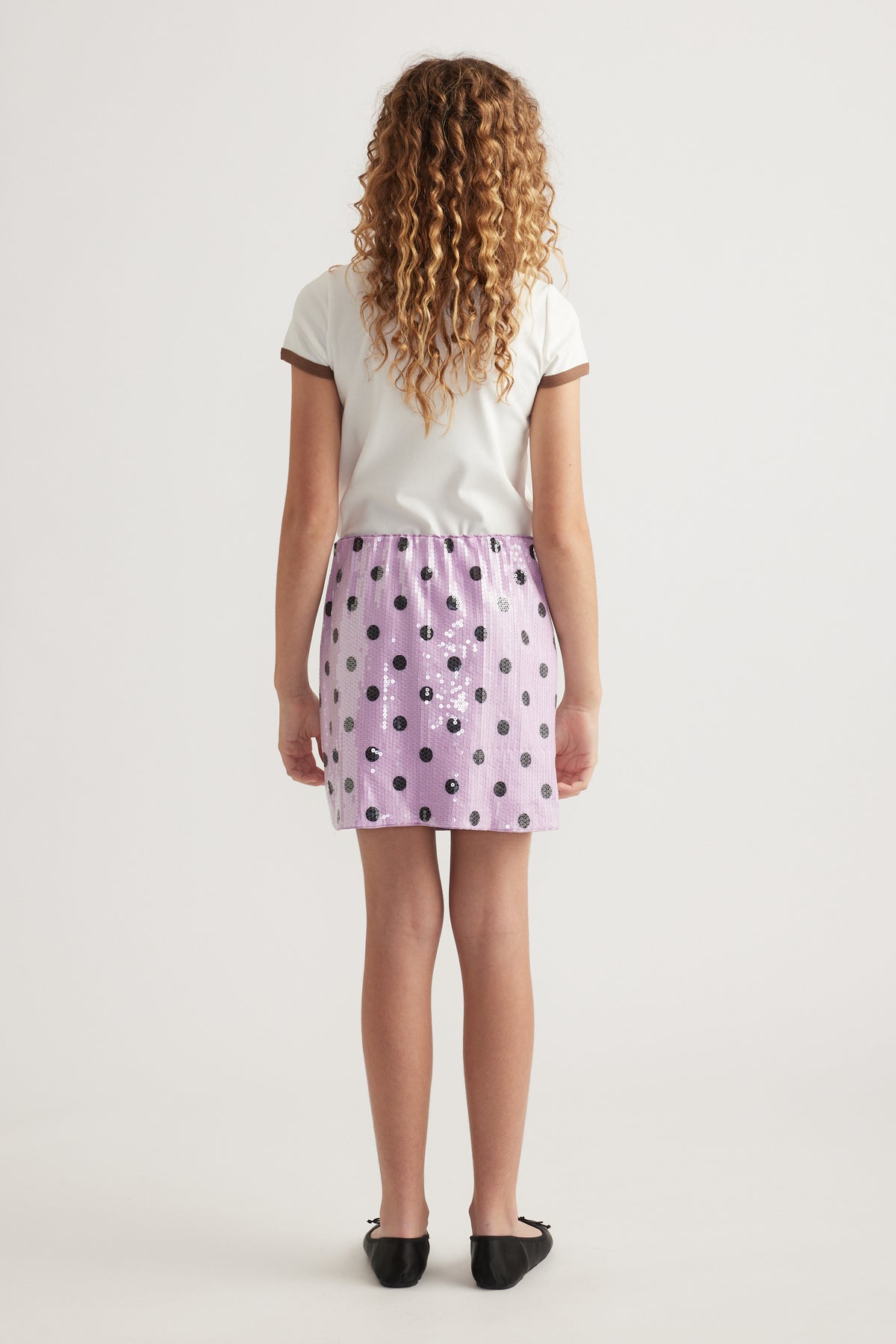Back view of a girl wearing a lavender sequin mini skirt with black spots, white tee and black flats