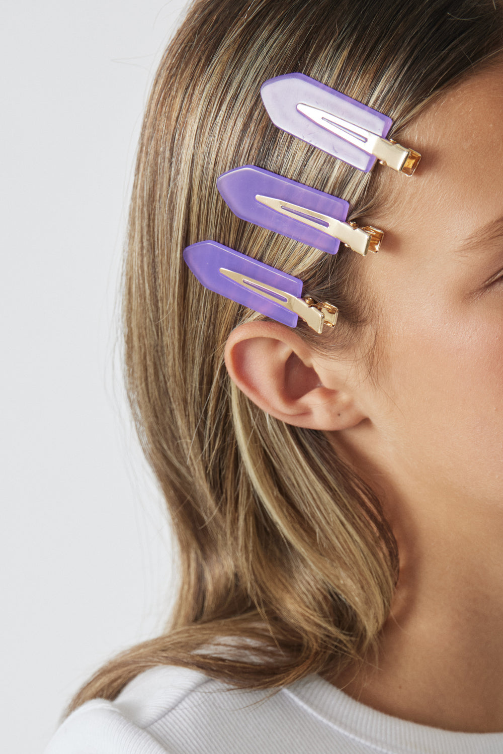 Three purple hair clips securing smooth light brown hair on side of head against plain background