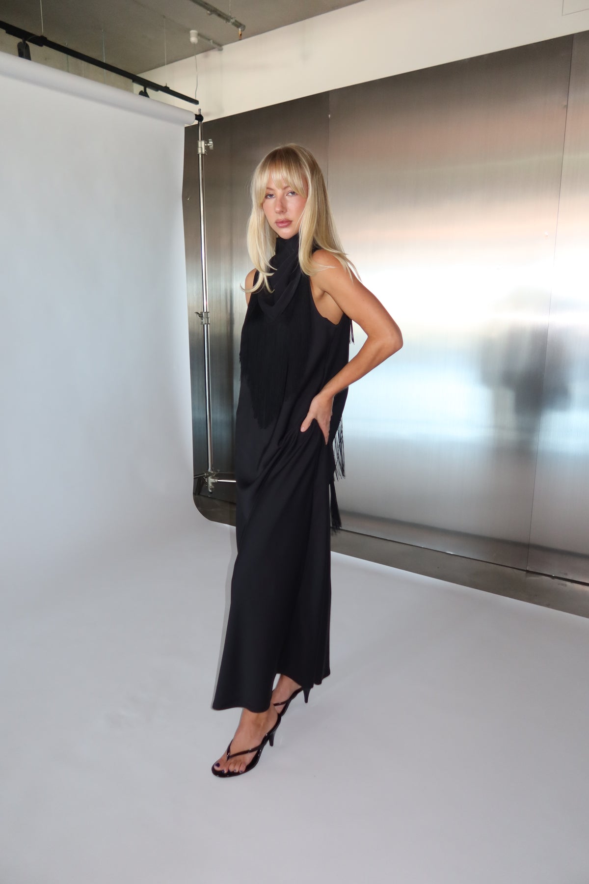 Woman wearing a black sleeveless maxi dress with a fringed waist scarf and strappy sandals