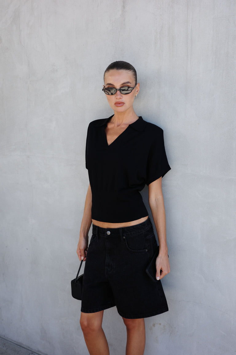 Woman in a black short sleeve polo knit top with black knee length shorts and a shoulder bag
