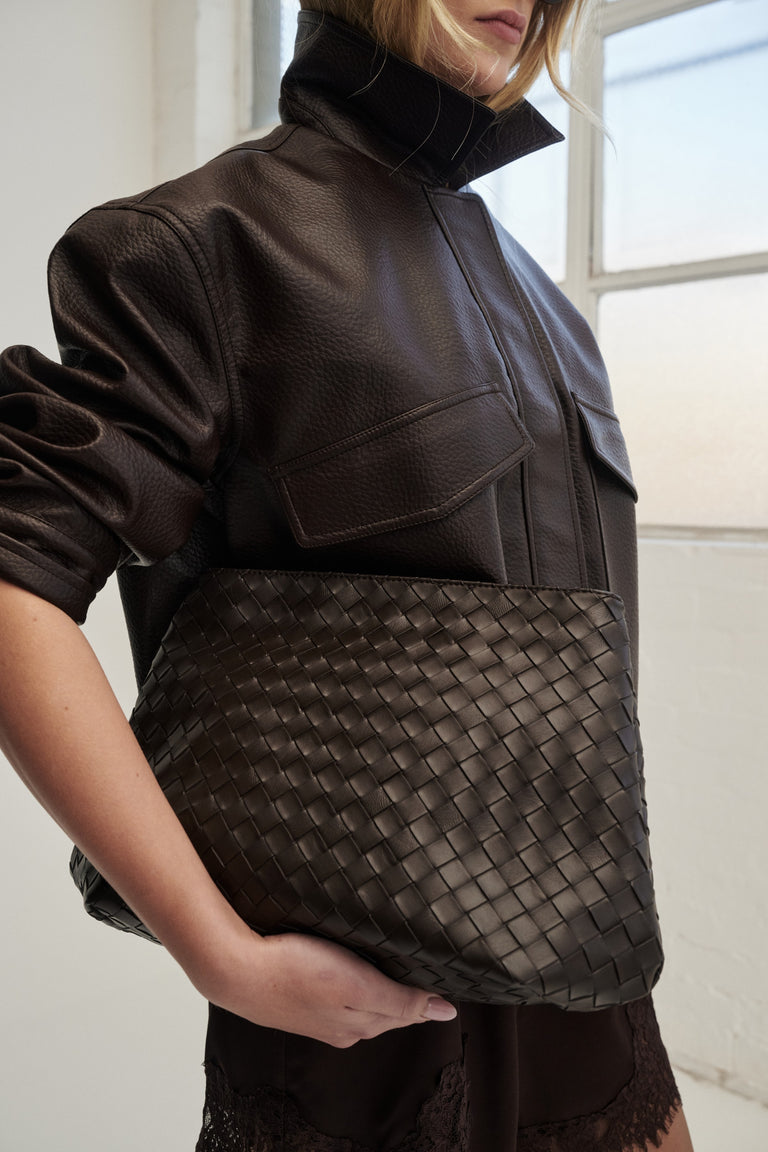 Woman holding a dark brown woven clutch beneath a black leather jacket