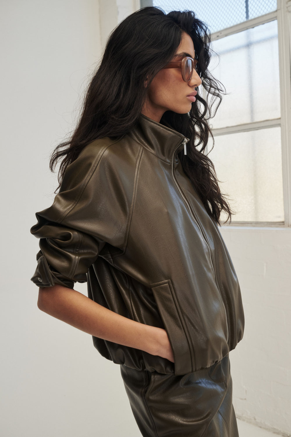 Woman wearing a dark brown faux leather bomber jacket with matching trousers and sunglasses