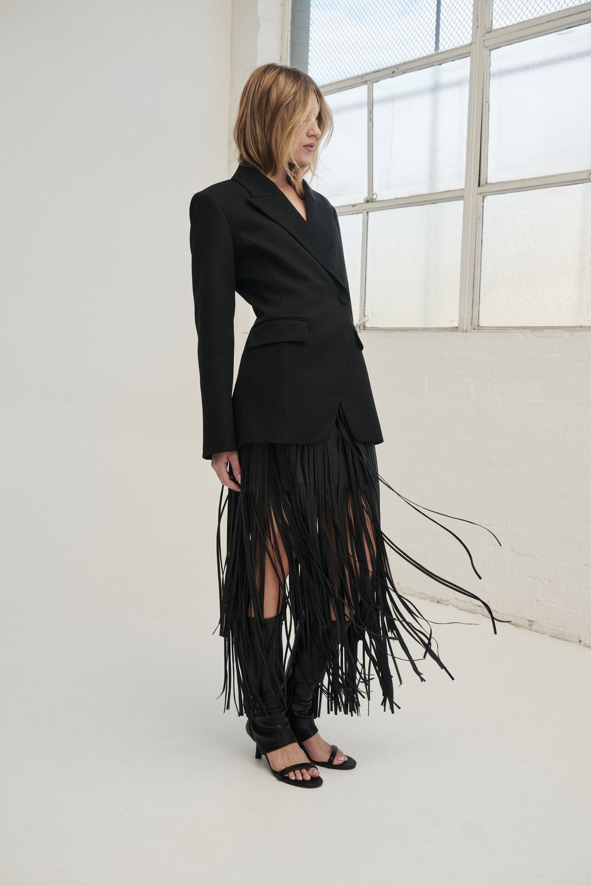 Woman wearing a black faux leather skirt with tiered fringe, styled with a tailored black top and flat sandals