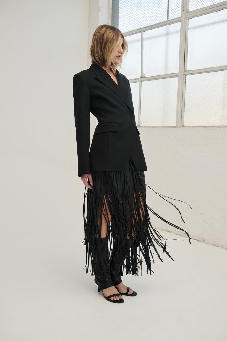 Woman wearing a black faux leather skirt with tiered fringe, styled with a tailored black top and flat sandals