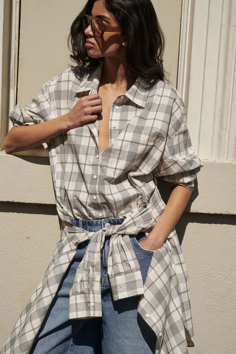 A woman wearing a light gray and white plaid button-down shirt tied at the waist over blue jeans, standing against a beige wall.