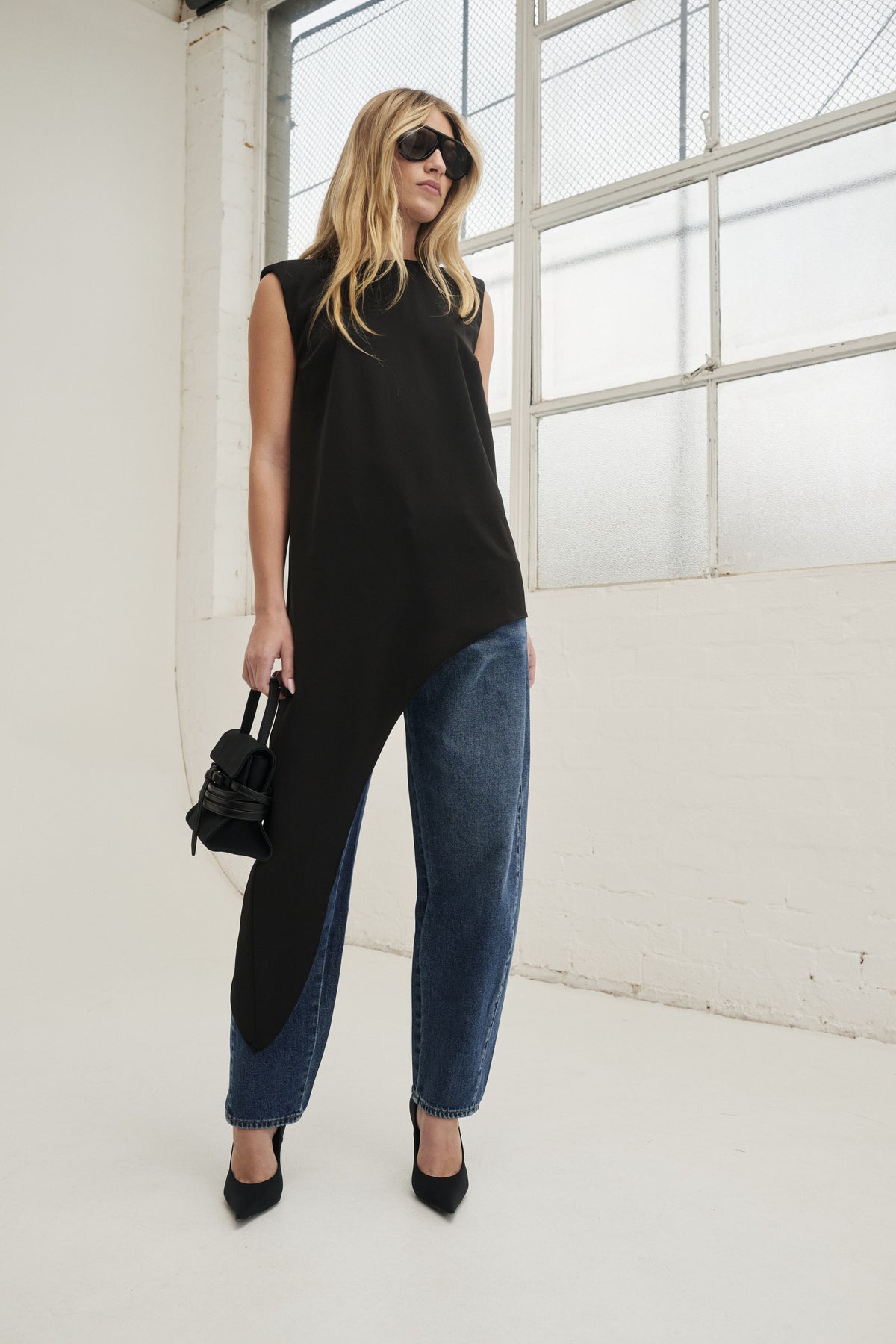 Woman in a black sleeveless draped top with an asymmetric hem, blue jeans and black heels
