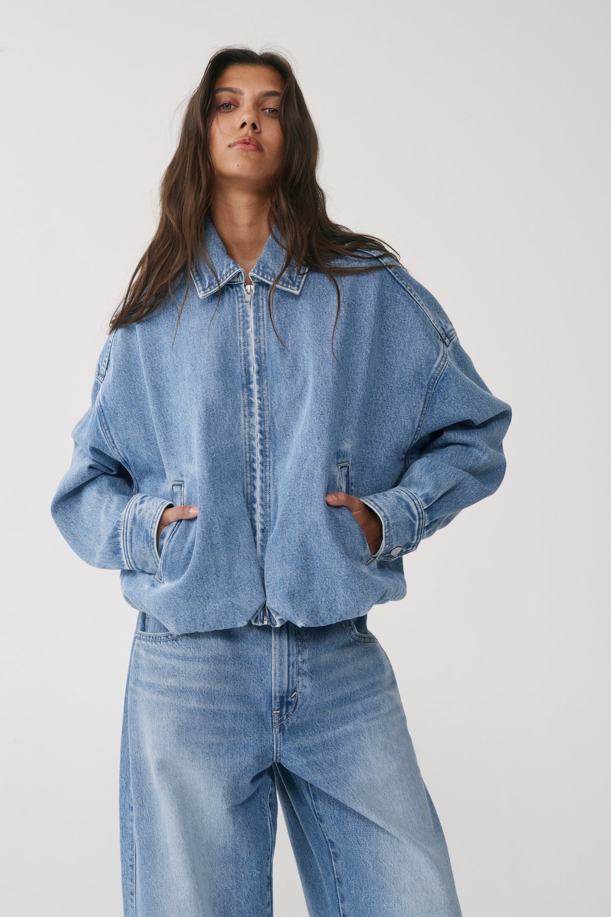 Woman wearing a light blue denim bomber jacket with a collared zip front and gathered hem