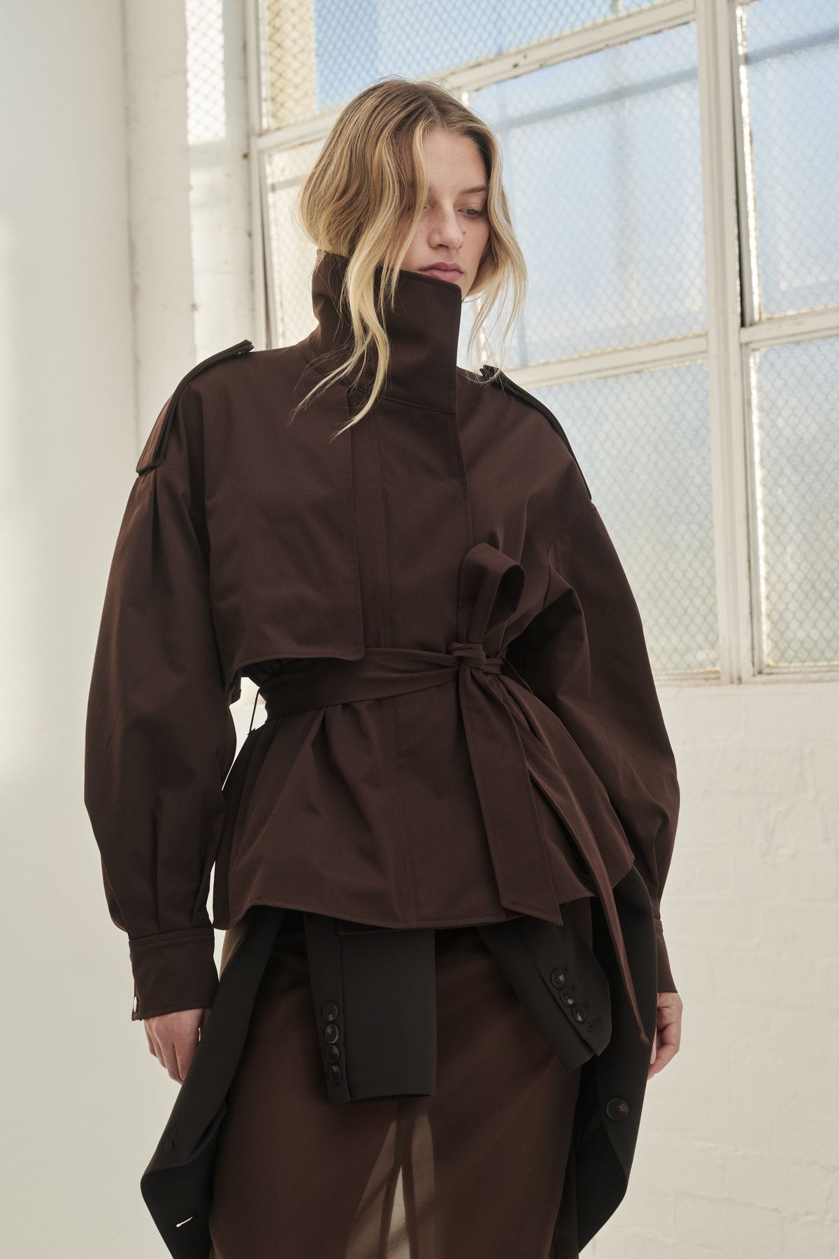Woman wearing a dark brown cropped trench coat with a high collar, shoulder epaulettes and tie waist