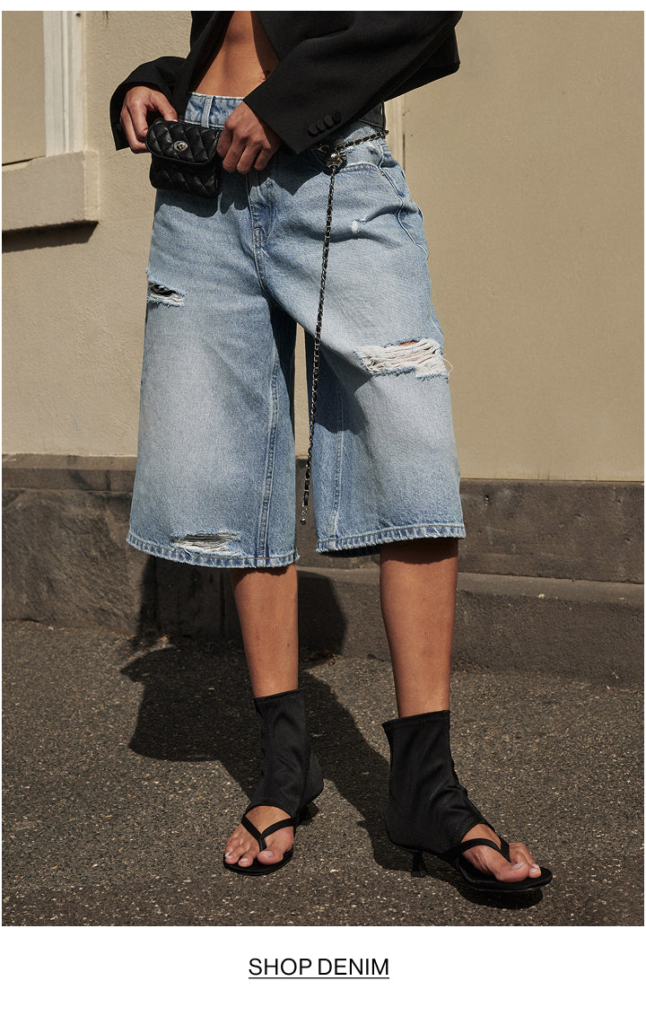 A person wearing light-wash distressed denim culottes with ripped details, styled with a black blazer, black ankle boots, and black sandals, standing outdoors against a beige wall.