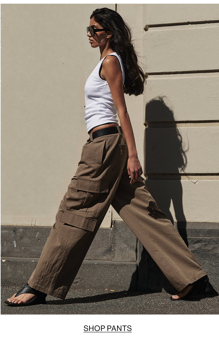 A woman wearing wide-leg brown cargo pants, a white tank top, and black sandals walks outdoors against a beige wall. The text 'SHOP PANTS' appears at the bottom of the image.