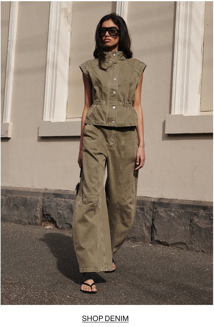 A woman wearing a matching olive green denim set consisting of a sleeveless button-front top and wide-leg pants, standing outdoors in front of a beige building wall. The text 'SHOP DENIM' is printed at the bottom of the image.
