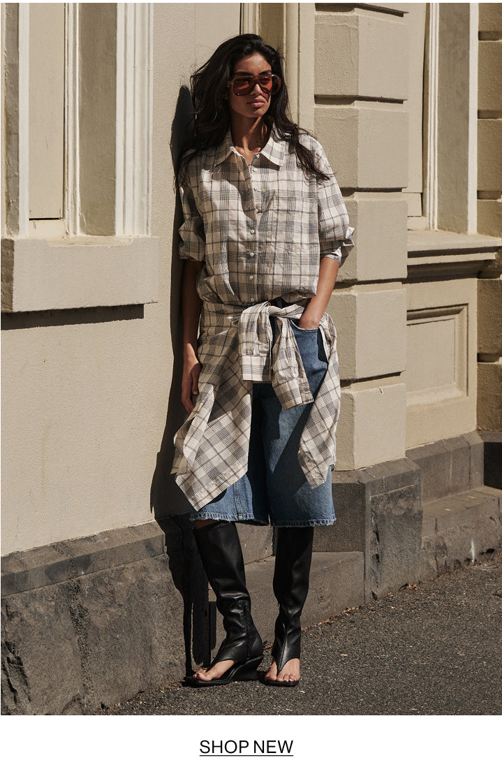 A woman stands outdoors against a beige stone building, wearing a beige and gray plaid button-up shirt tied at the waist over blue denim shorts, black knee-high boots, and dark sunglasses. A white banner at the bottom reads 'SHOP NEW'.