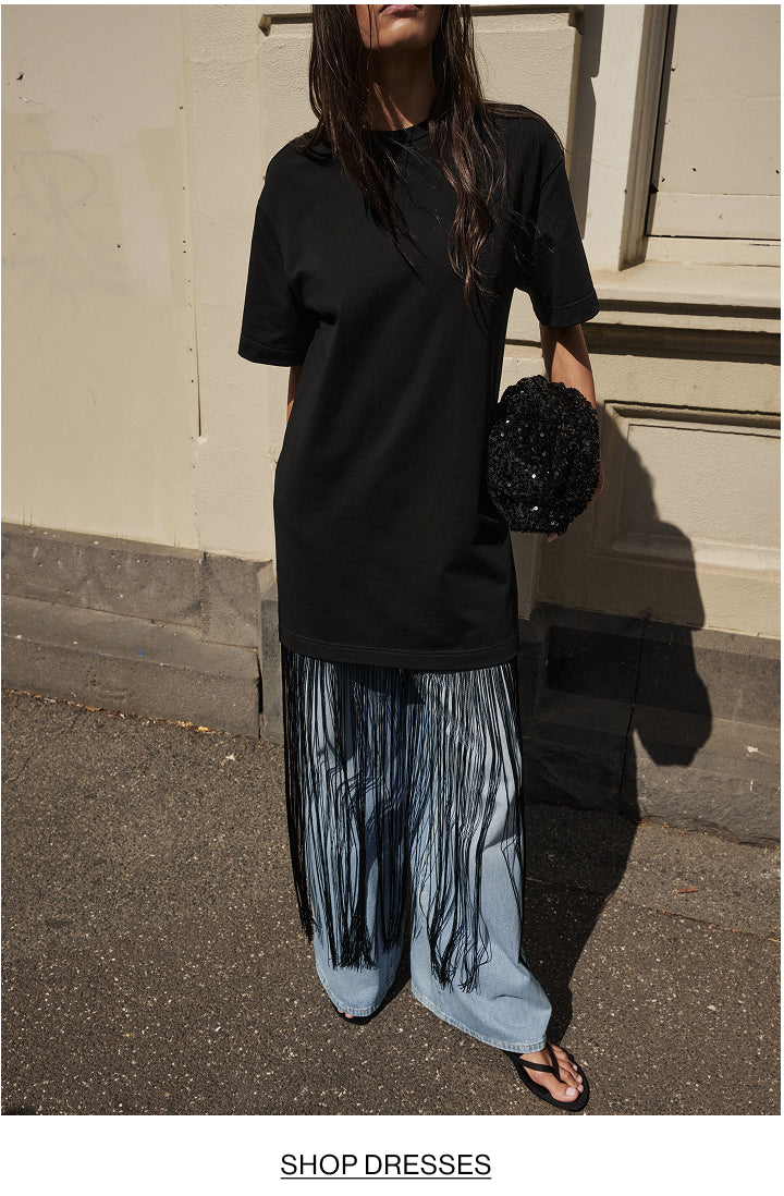 A woman wearing an oversized black short-sleeve top and wide-leg light blue jeans with long fringe, holding a black sequined clutch bag, standing outdoors against a beige wall.