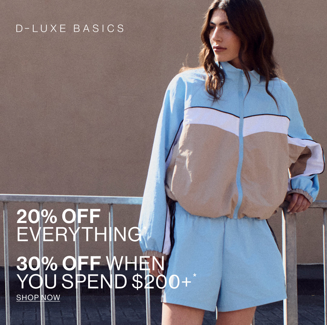 A woman models a light blue and beige color-blocked windbreaker jacket with matching light blue shorts against a textured brown wall, with promotional text overlays for D-Luxe Basics.