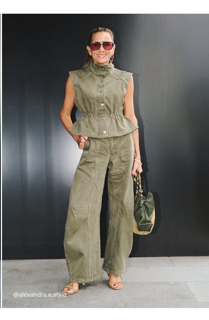 A woman modeling a matching olive green sleeveless button-front top and wide-leg pants set, styled with sunglasses, a green and beige handbag, and flat sandals against a dark studio backdrop.
