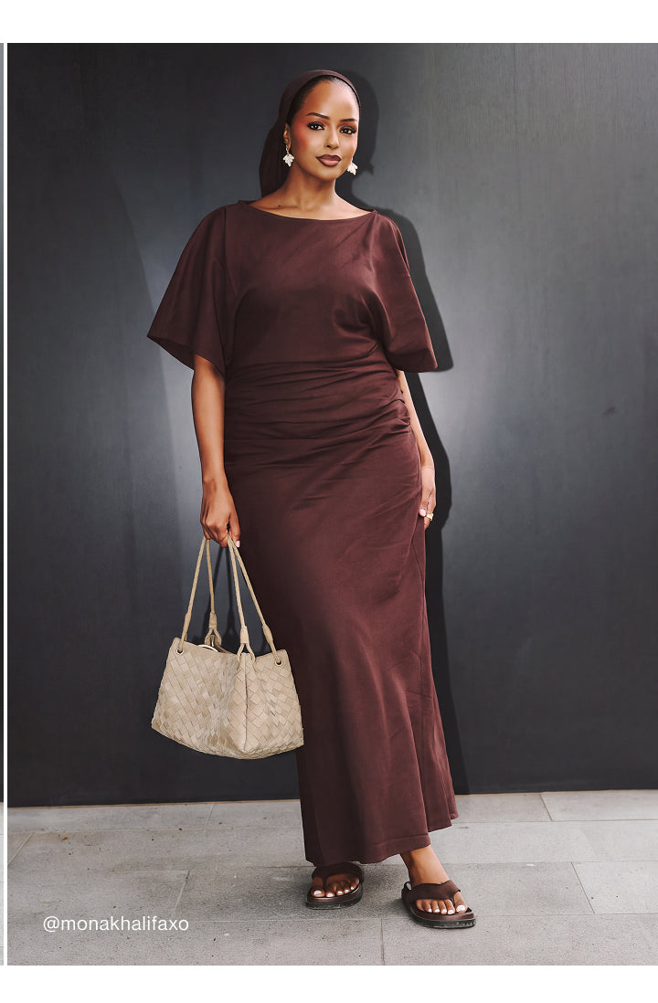 A woman modeling a dark brown, short-sleeved maxi dress with a gathered, ruched bodice and flowing skirt, holding a beige textured tote bag against a dark gray wall.