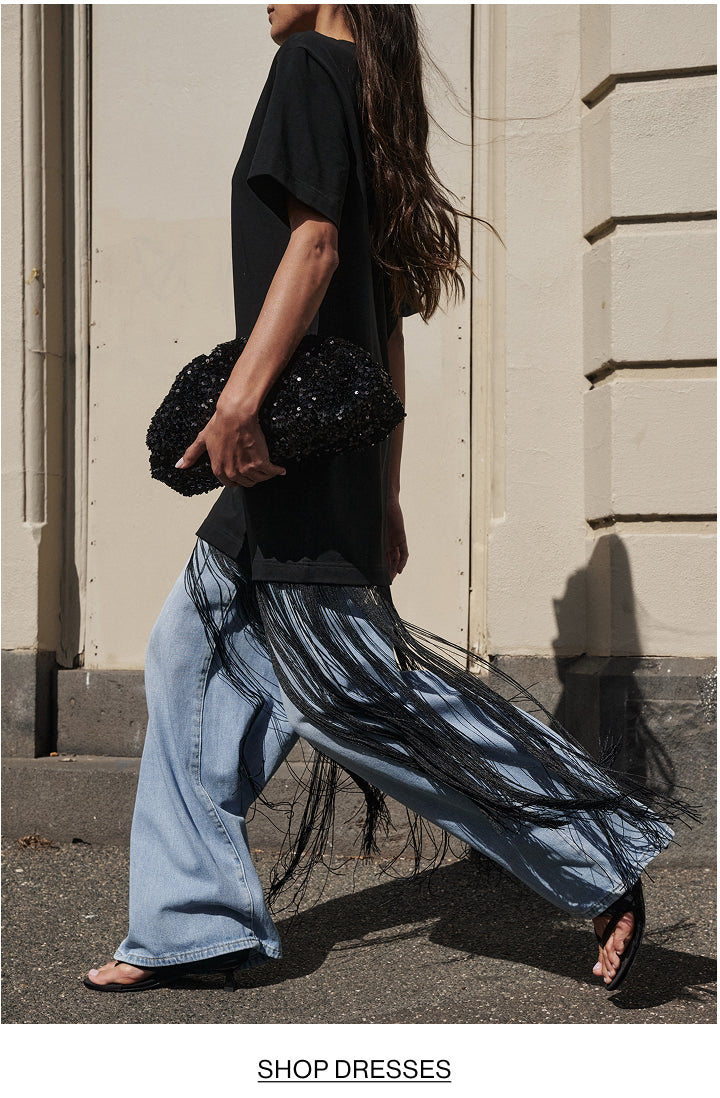 A woman walking outdoors wearing a loose black oversized T-shirt, very wide-leg light blue jeans with long black fringe cascading down the front, black sandals, and holding a black beaded clutch bag.