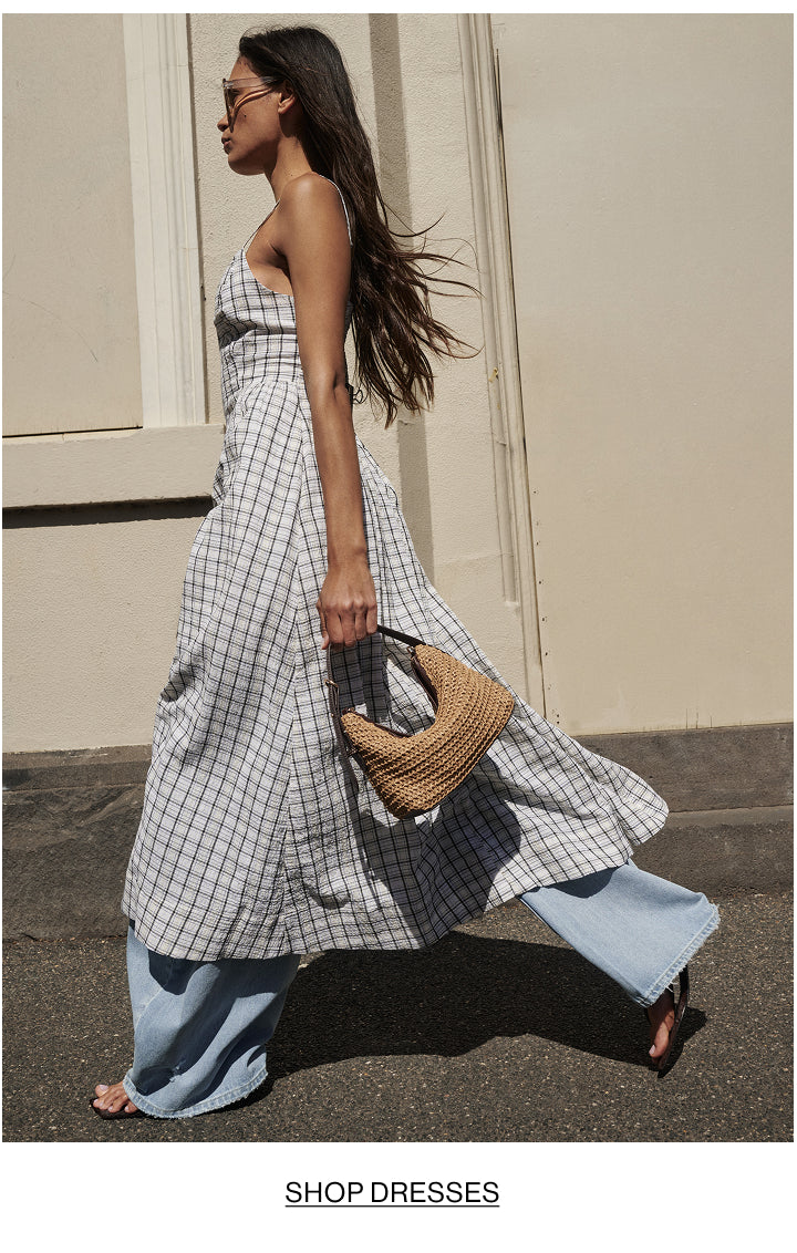 A woman walking outdoors in a sleeveless white and gray gingham maxi dress, styled with wide-leg light blue jeans and carrying a woven straw handbag.