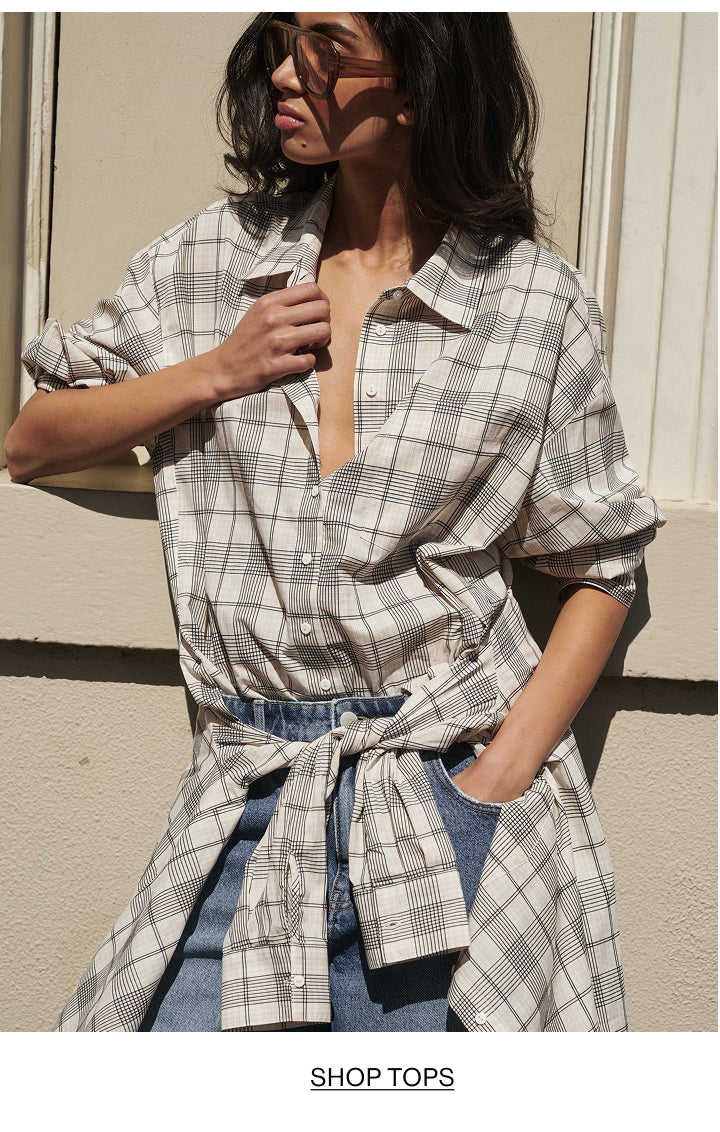 A woman wearing an oversized white and gray plaid button-up shirt tied at the waist over blue jeans, standing against a beige wall.