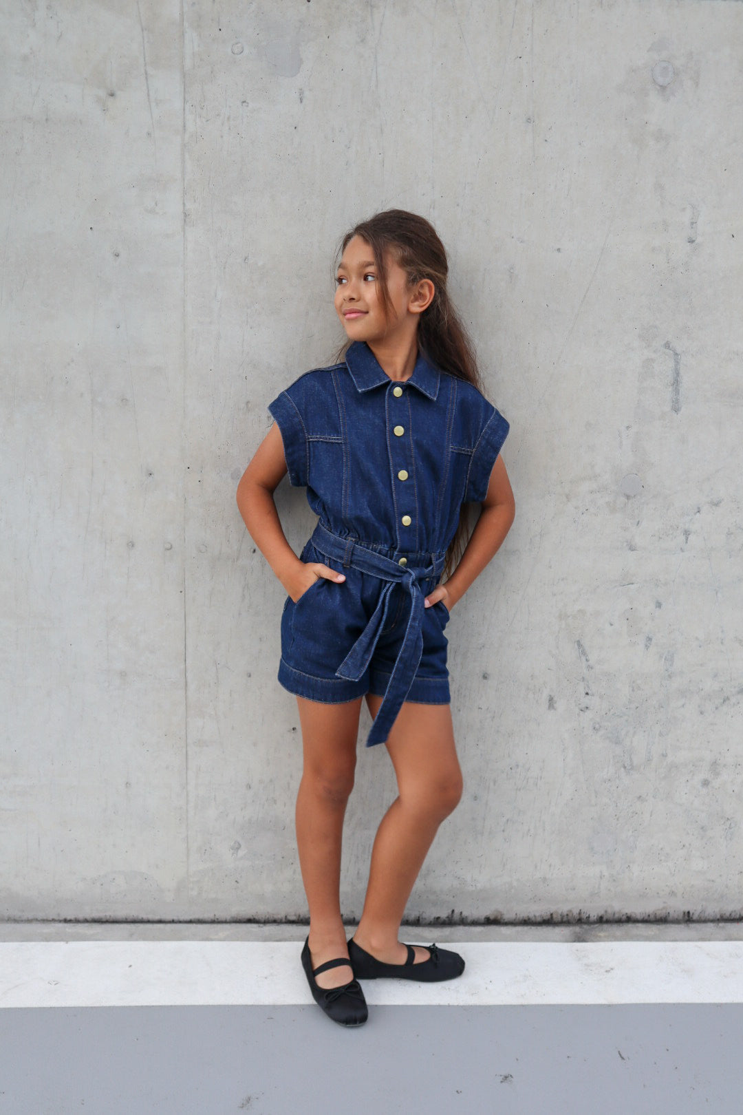 Young girl wearing a belted denim romper paired with black satin ballet flats, standing against concrete wall background