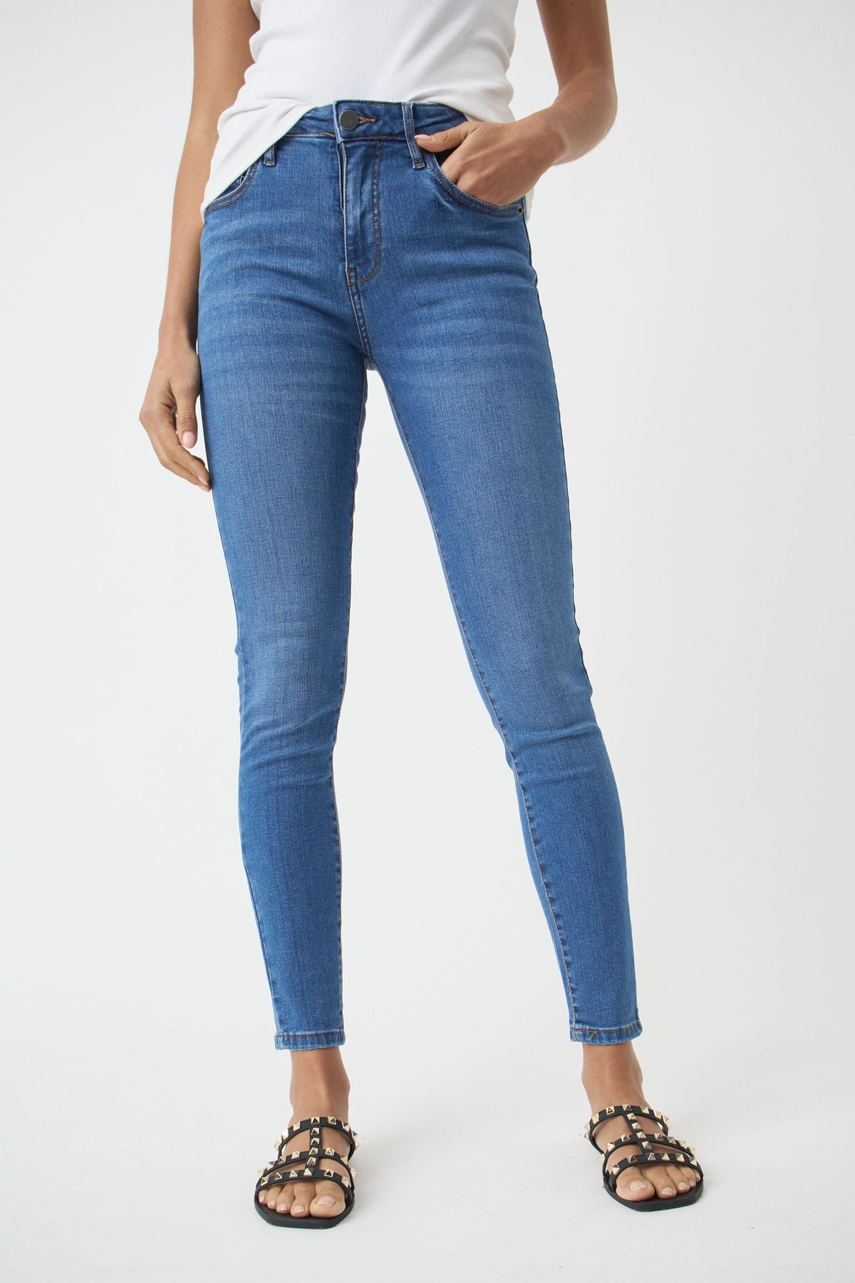 Woman wearing mid-blue skinny jeans with a high waist, paired with a white top and studded black sandals, standing relaxed