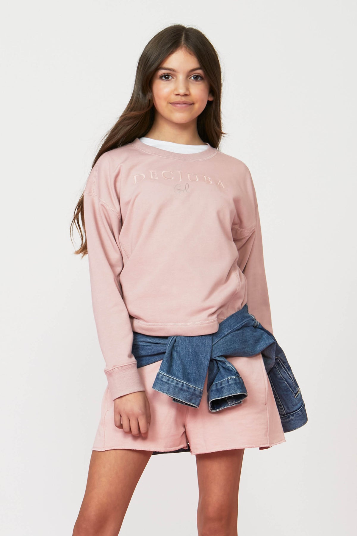 Girl wearing a pink crew neck sweatshirt with matching shorts and a denim jacket tied at the waist